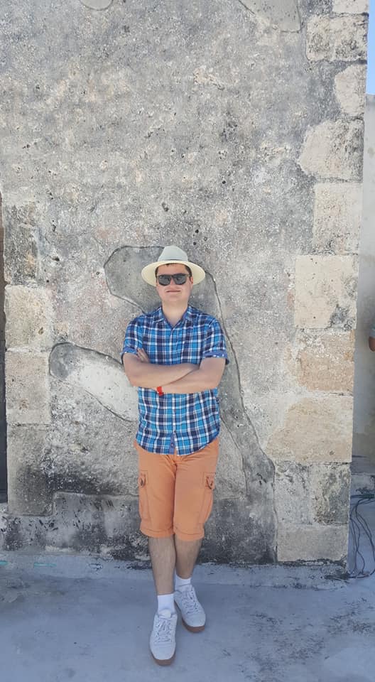 Young man wearing a plaid shirt, orange shorts, white sneakers, and a straw hat leaning against the wall.