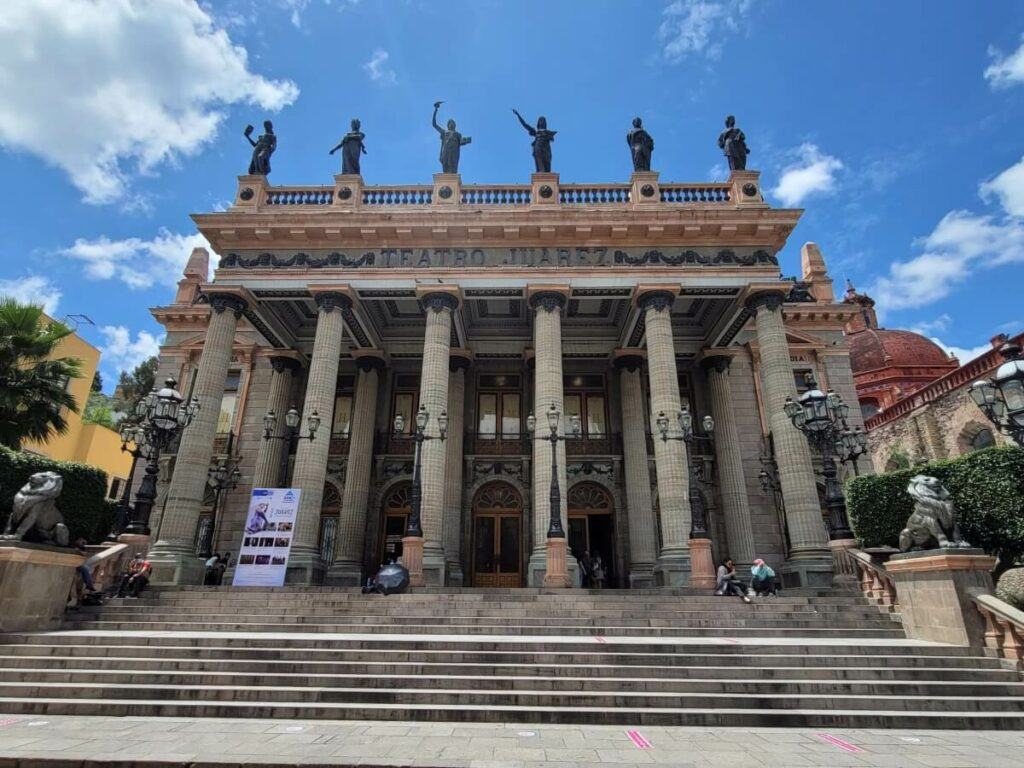 Entrance of a theater with doric columns and statues on the roof.