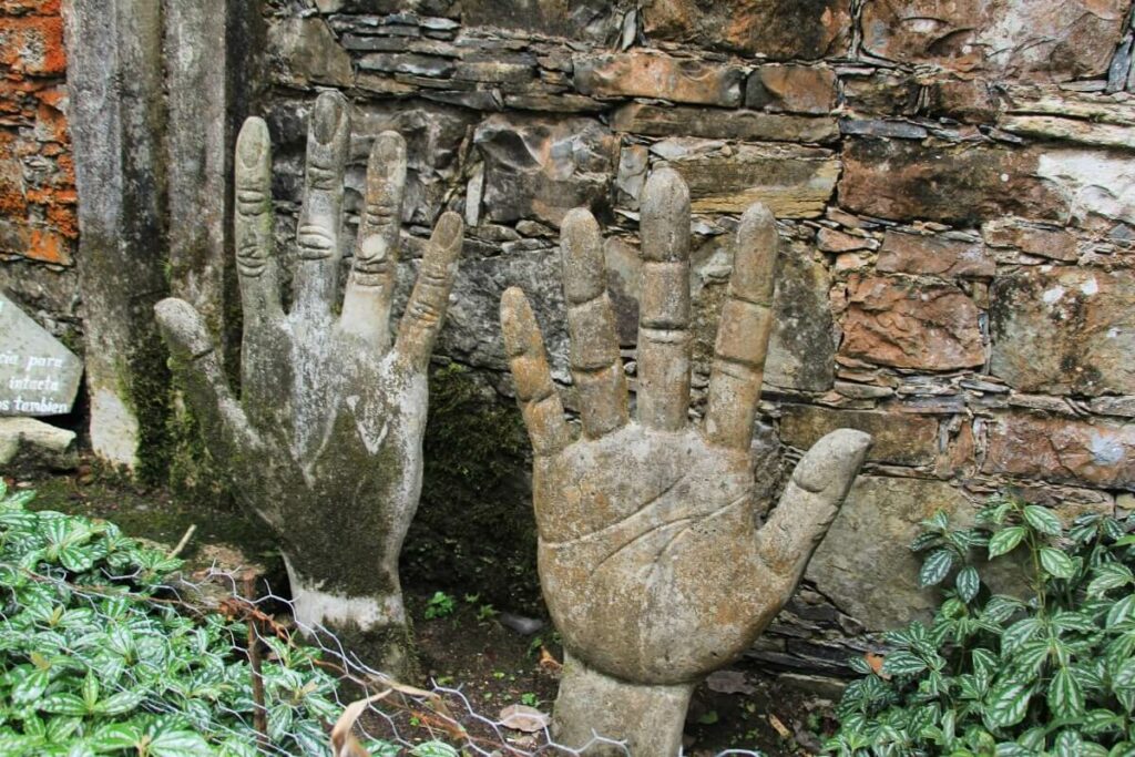 Two sculptures shaped like hands surrounded by plants and a stone wall in the back.