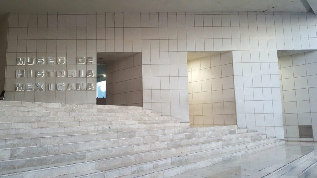 The facade of a museum consisting of pearl white tiles and the letteres reading Museo de Historia Mexicana.