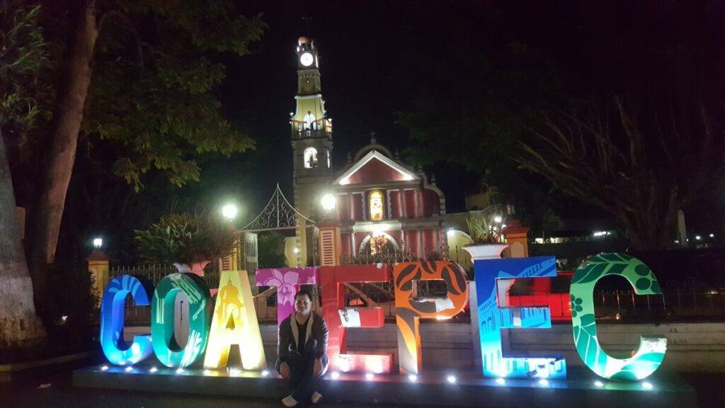 A red and yellow church lit up at night with the letters Coatepec in front of it.