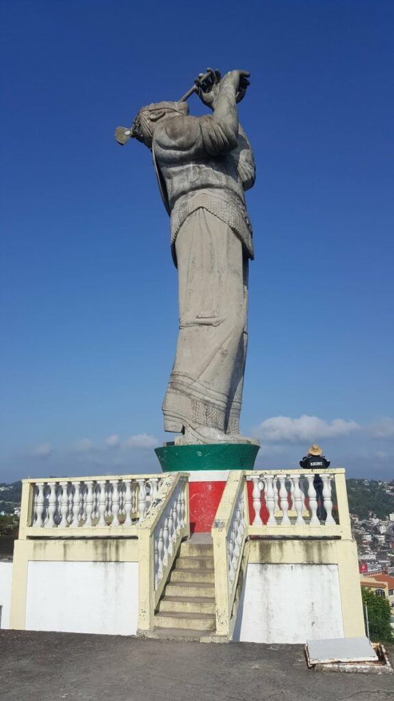 A monument of a man dressed in Totonac clothing and playing the flute.